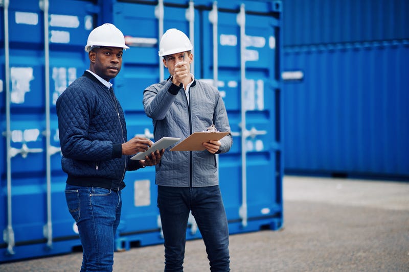 Two engineers in white hardhats carrying clipboards and a tablet stand in front of a blue shipping container